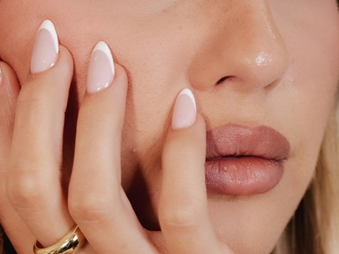 A close-up of soft pink almond nails resting against the cheek beside glossy nude lips